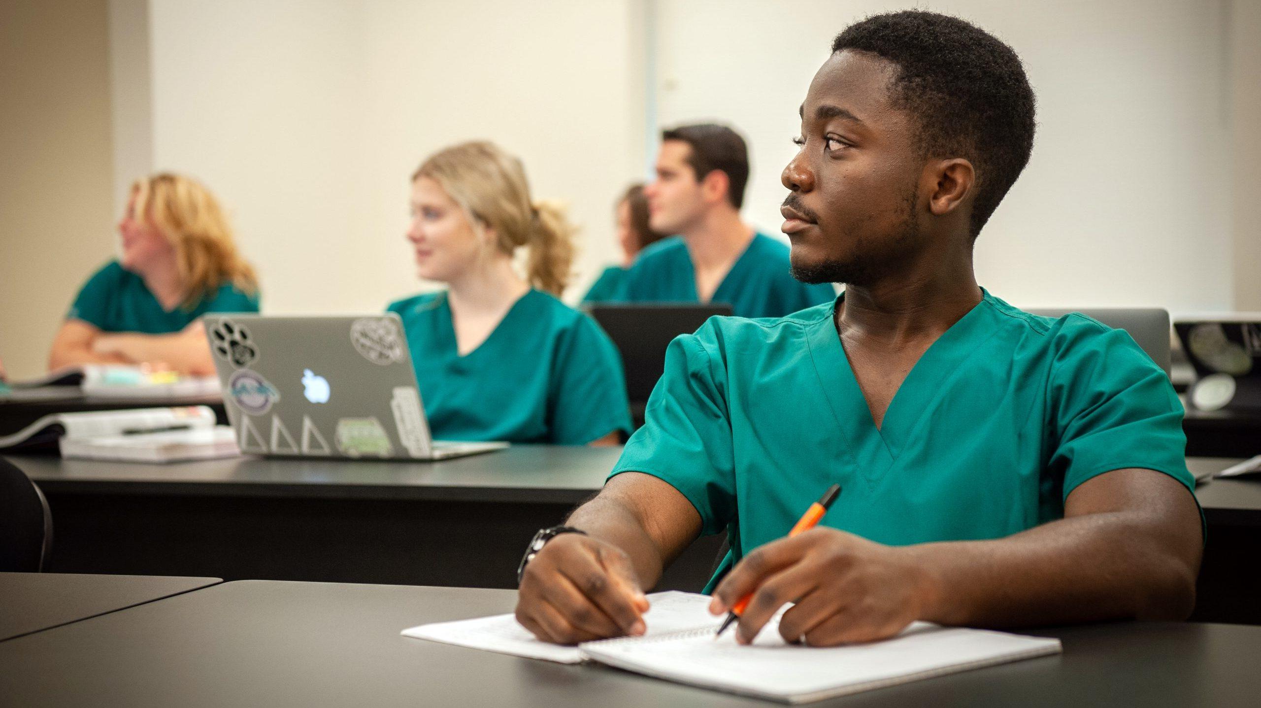 Student sitting at a table in class, taking notes during lecture, with four other students behind him.